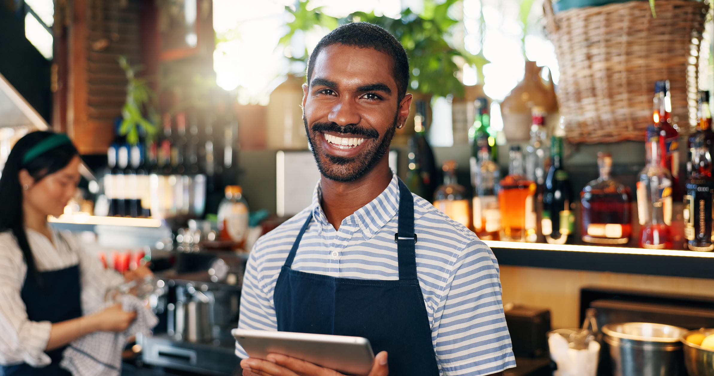 Male bartender smiling and holding a tablet, while a female bartender dries glass in the background