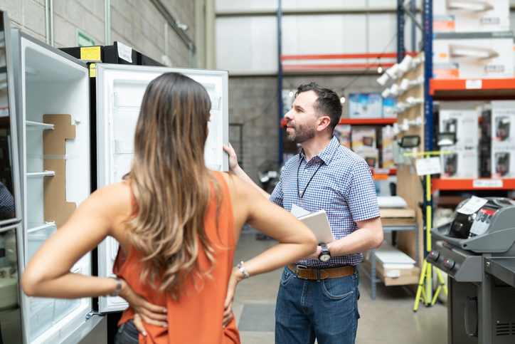 Male employee showing while explaining about refrigerator to female customer standing at warehouse store