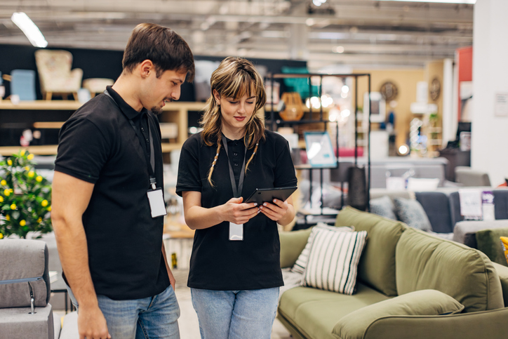 A saleswoman in a department store uses a digital tablet while organizing furniture in a store with her male colleague