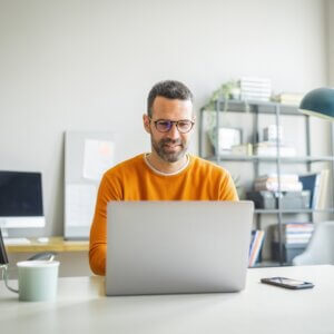 Businessman wearing casual clothing and eyeglasses sitting at his desk and using laptop in the office.